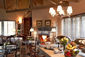 A dining area with a fireplace, wooden chairs, a table set with plates and a fruit bowl, and a chandelier.