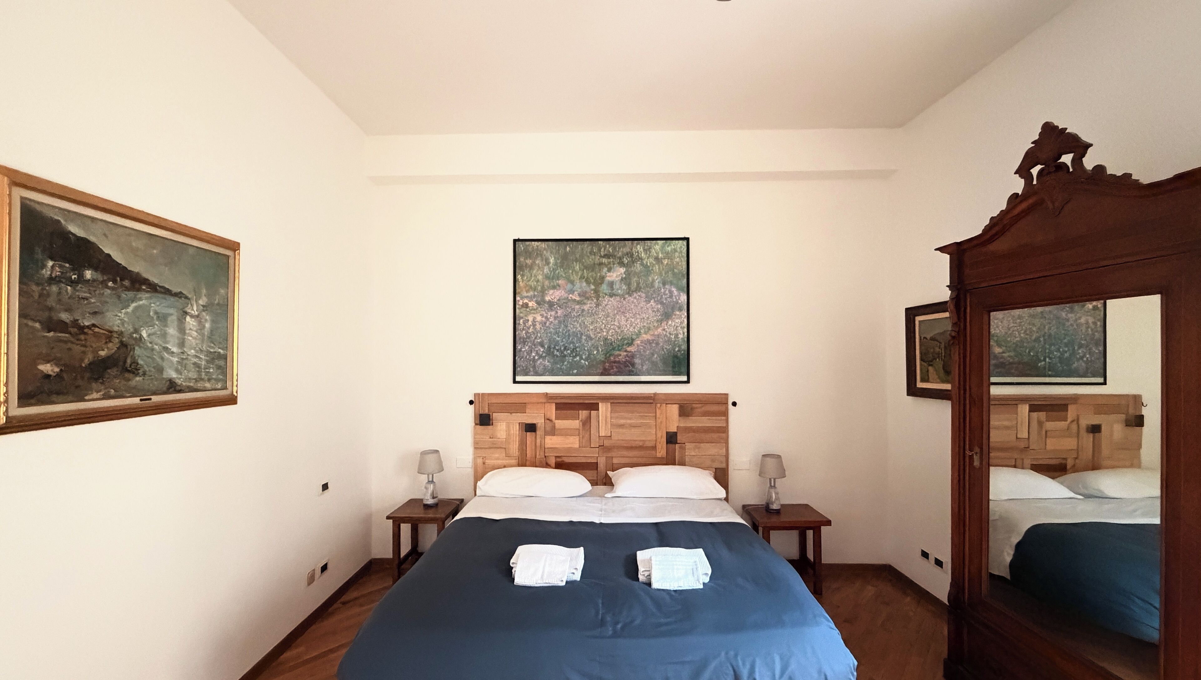 A bedroom with a wooden headboard, two bedside tables with lamps, a bed with blue and white bedding, and two paintings on the walls.
