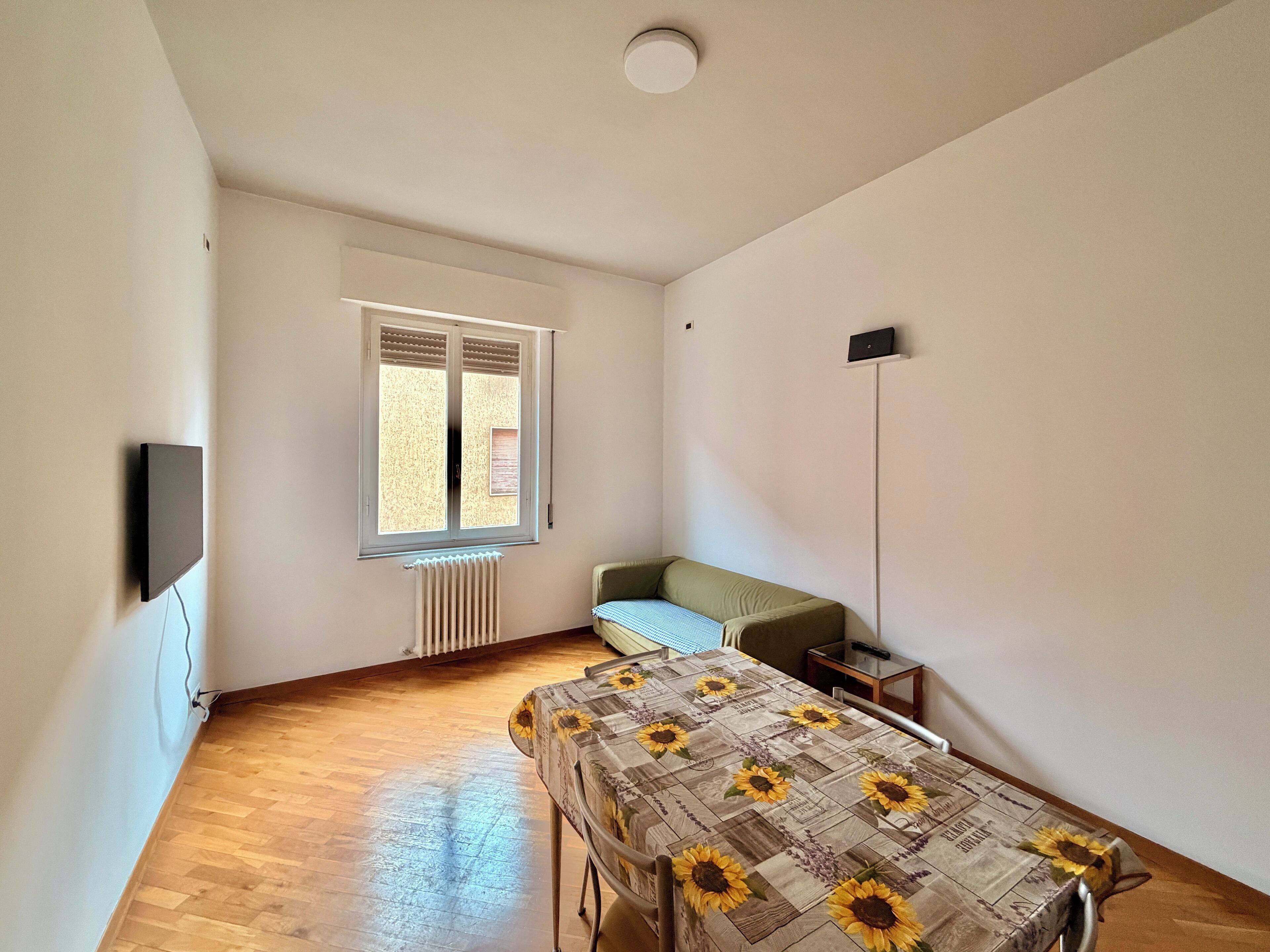Ein Zimmer mit einem Bett, einem Heizkörper, einem Fenster mit Jalousien, einer Stehlampe und einem an der Wand befestigten Fernseher.