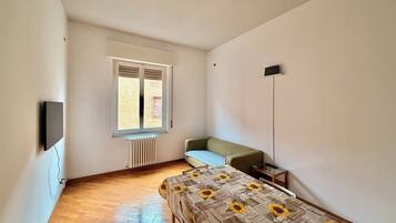 Ein Zimmer mit einem Bett, einem Heizkörper, einem Fenster mit Jalousien, einer Stehlampe und einem an der Wand befestigten Fernseher.