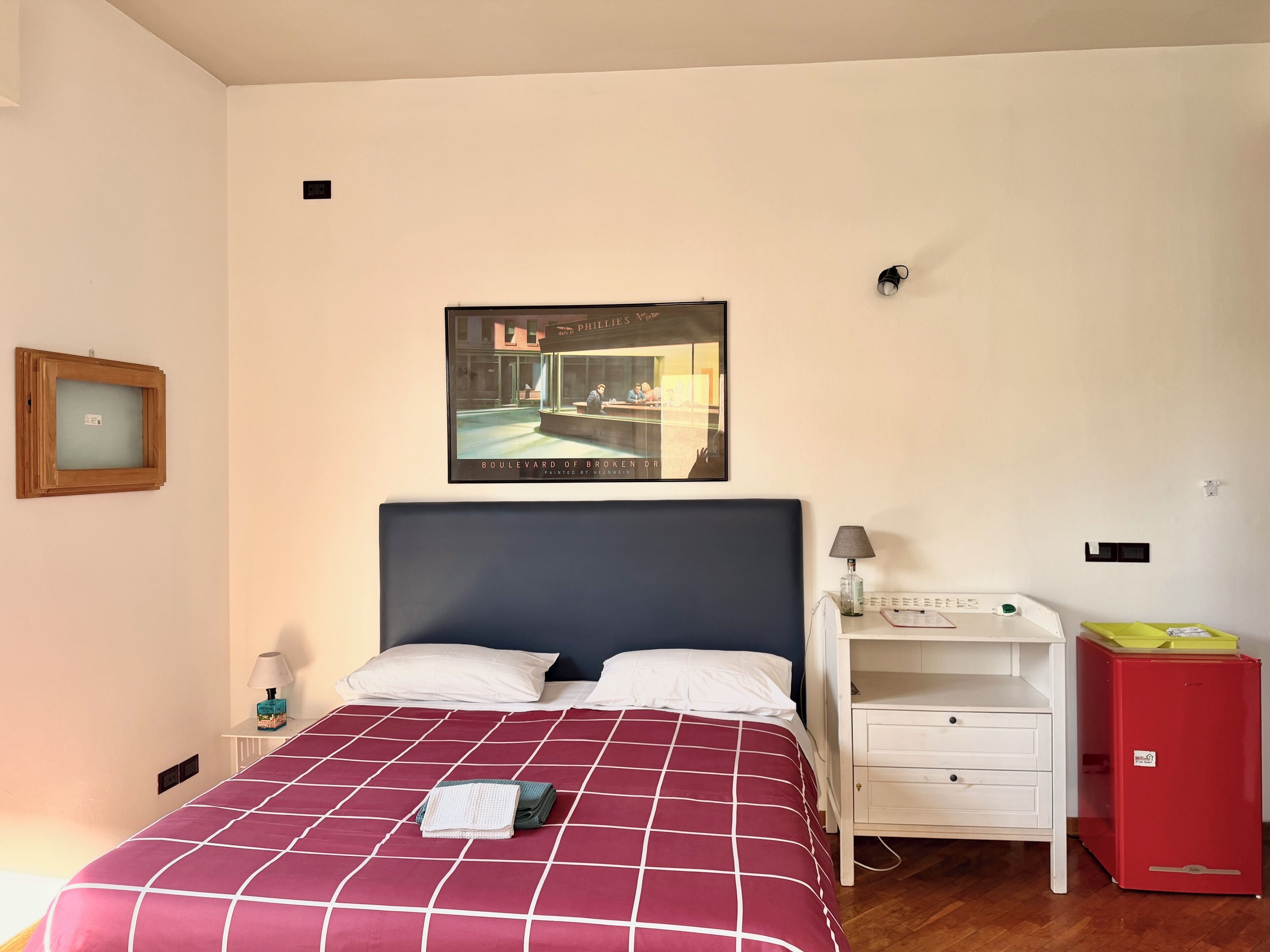 A hotel room with a bed, a nightstand, a desk, a red refrigerator, and a poster on the wall.