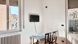 Una habitación con una mesa de comedor y sillas, un televisor montado, un radiador y una ventana con persianas.