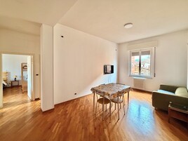 Amplia sala de estar con piso de madera, una mesa de comedor con sillas, un sofá y un televisor montado en la pared.