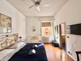 Un dormitorio con una cama, ventilador de techo, escritorio, silla, televisor y ventana con persianas.