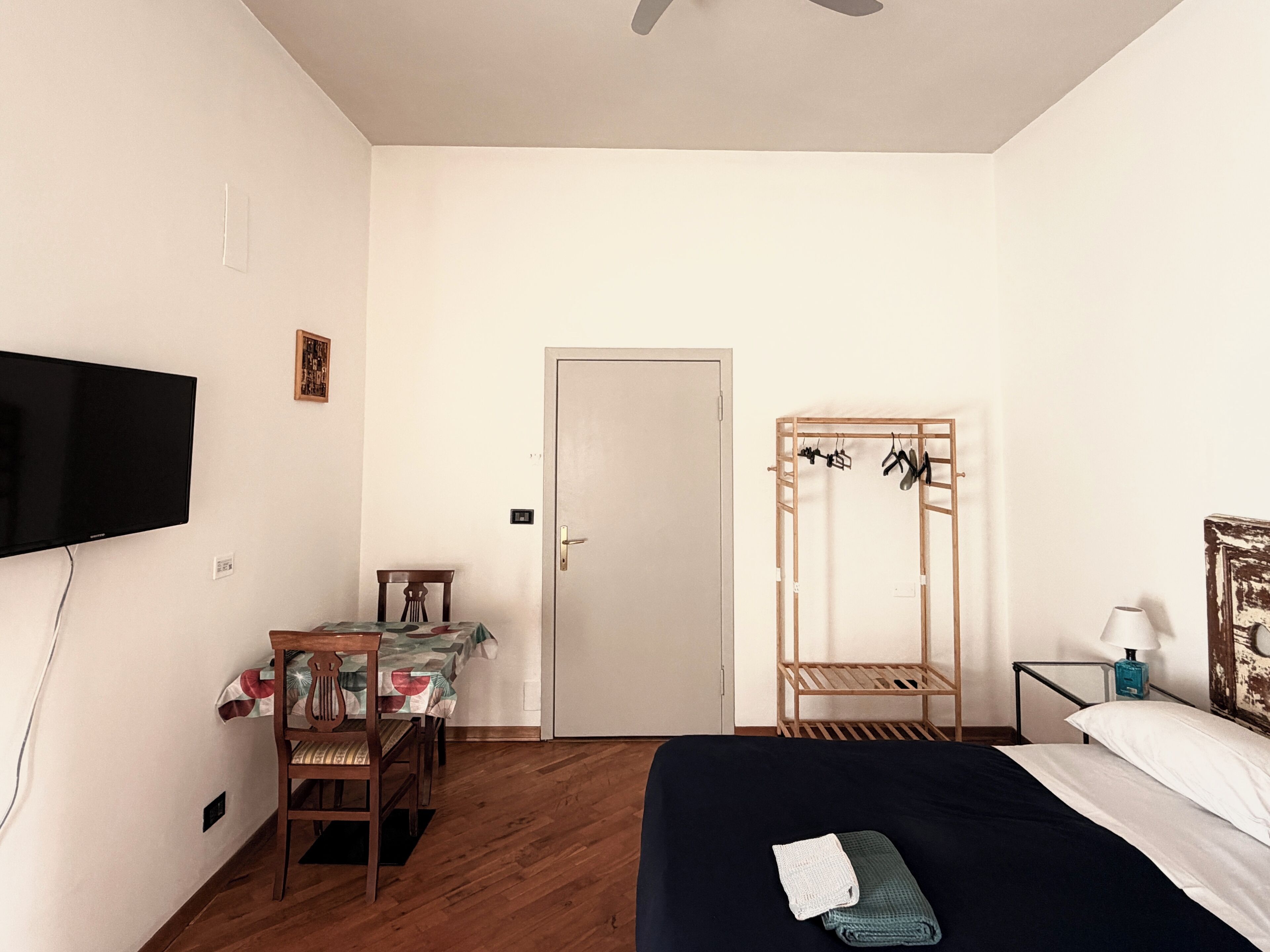 A bedroom with a bed, a TV mounted on the wall, a wooden chair, a table with a tablecloth, a door, and a wooden shelf with items on it.