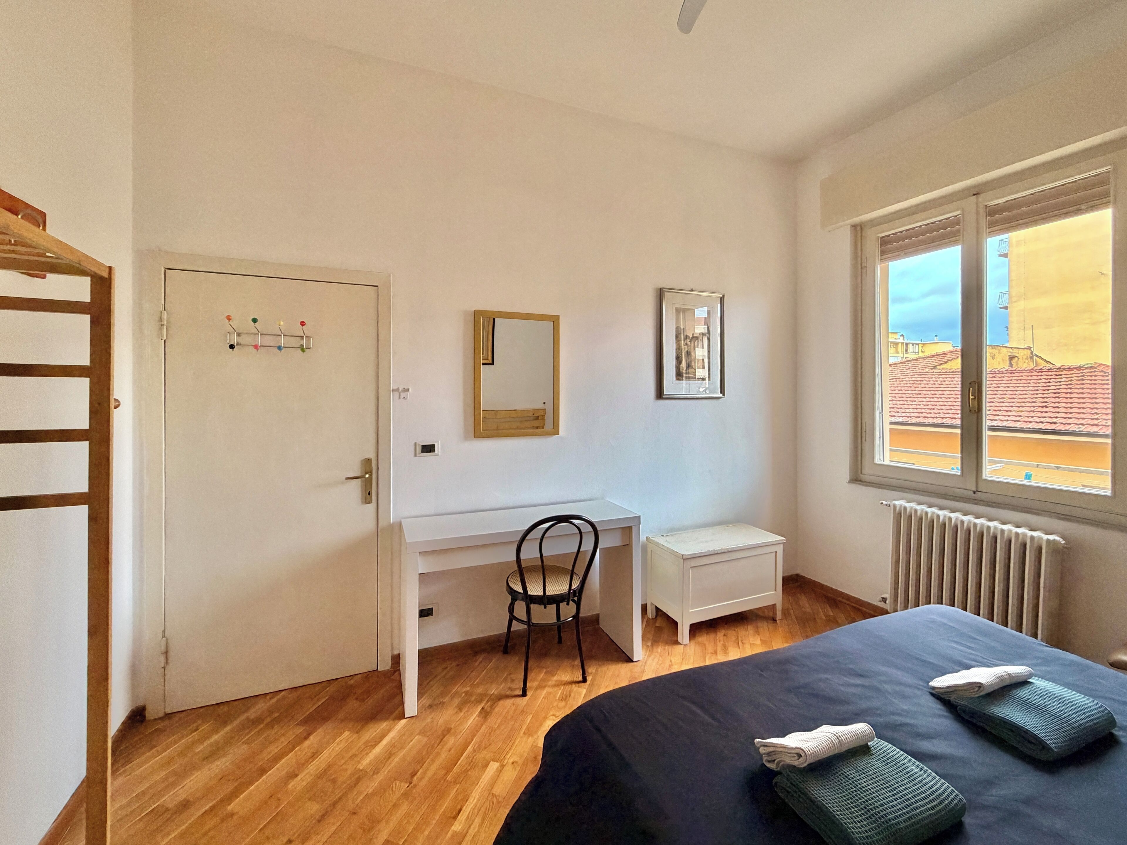 Un dormitorio con cama, escritorio, silla, radiador y ventana con vista a edificios.