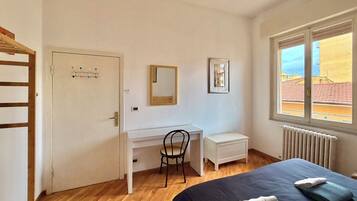 Un dormitorio con cama, escritorio, silla, radiador y ventana con vista a edificios.
