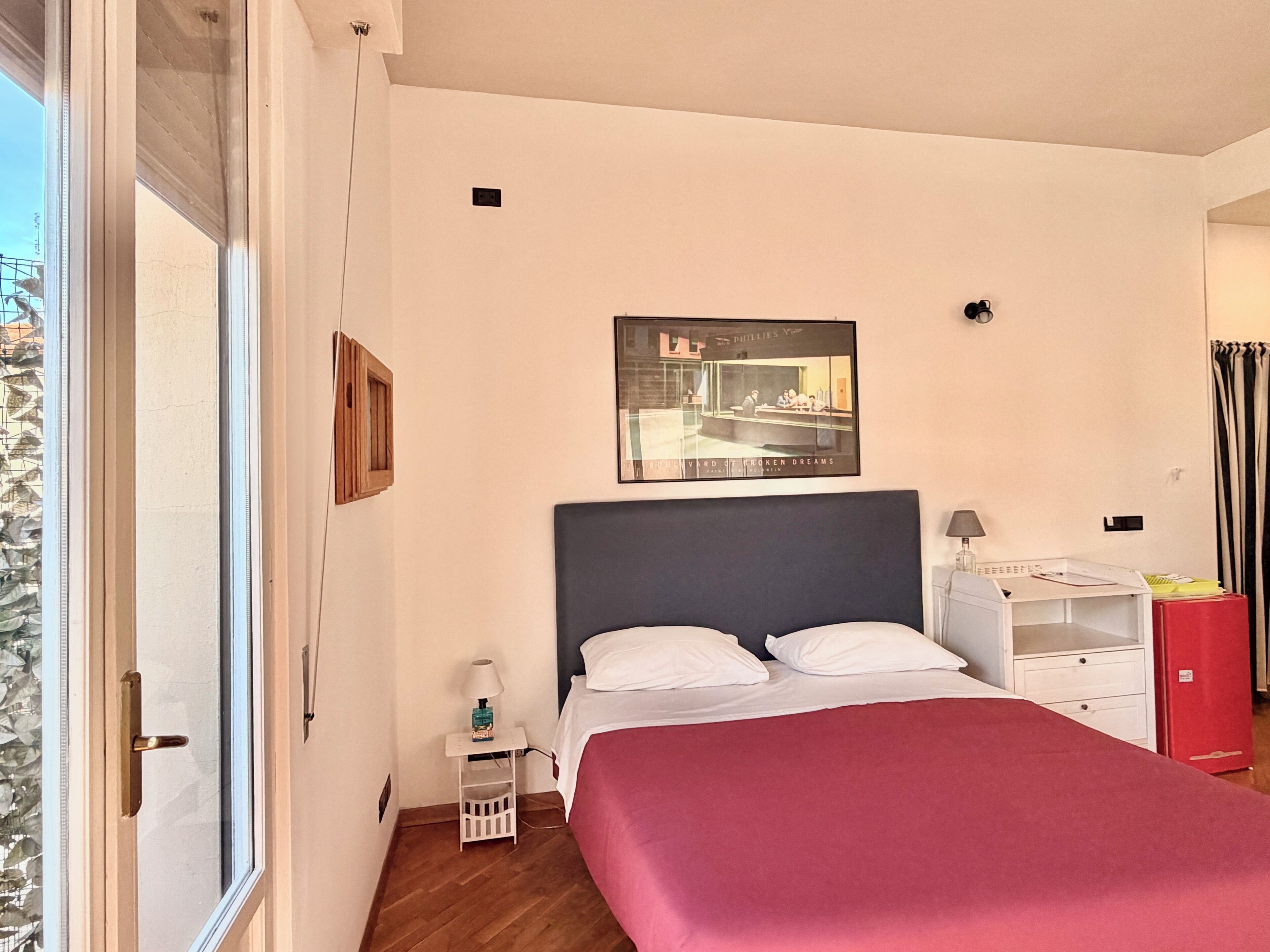 A bedroom with a bed, a nightstand, a lamp, a red cabinet, and a poster on the wall.