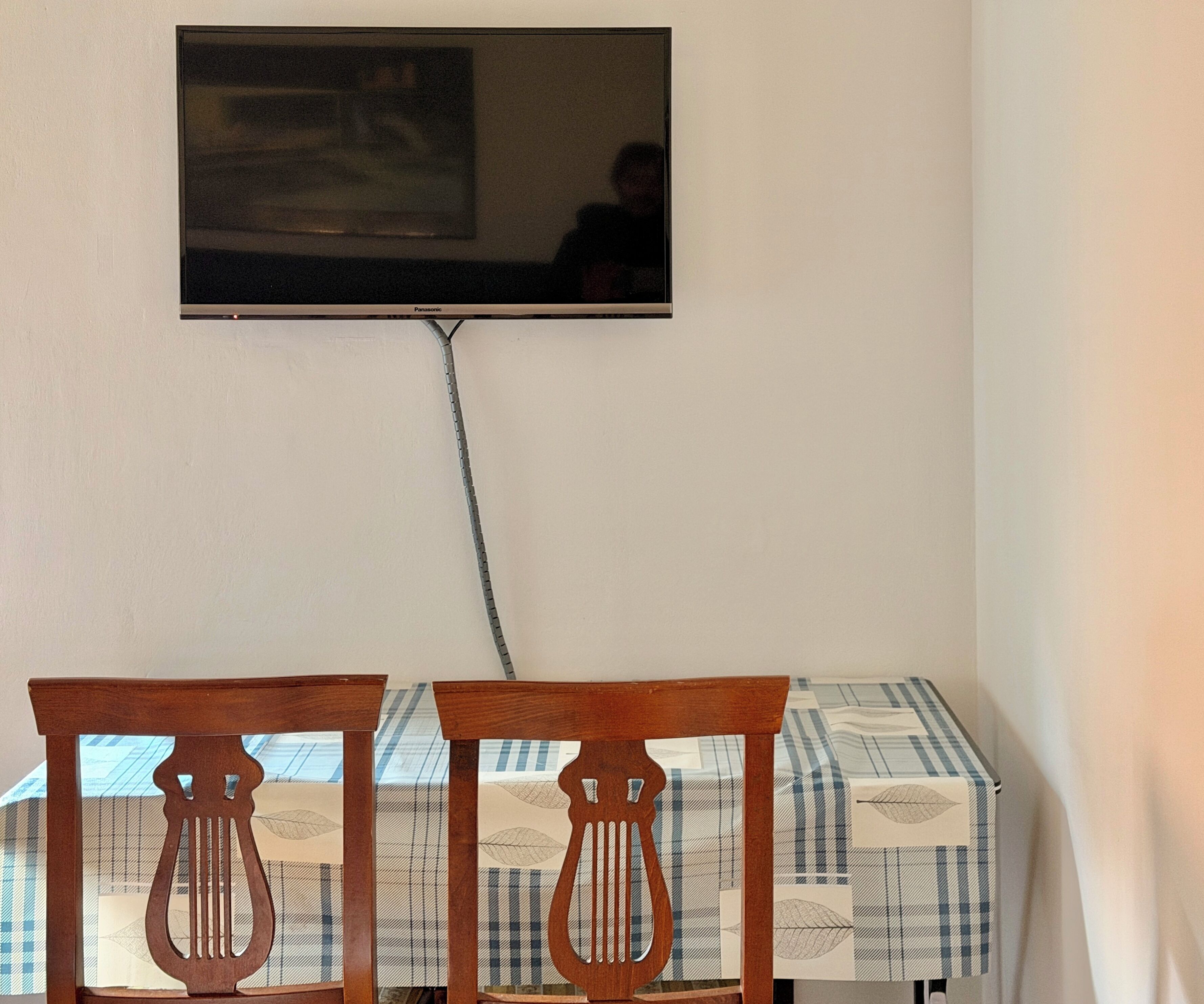Een eethoek met twee houten stoelen, een tafel met geruit tafelkleed en een aan de muur bevestigde televisie.