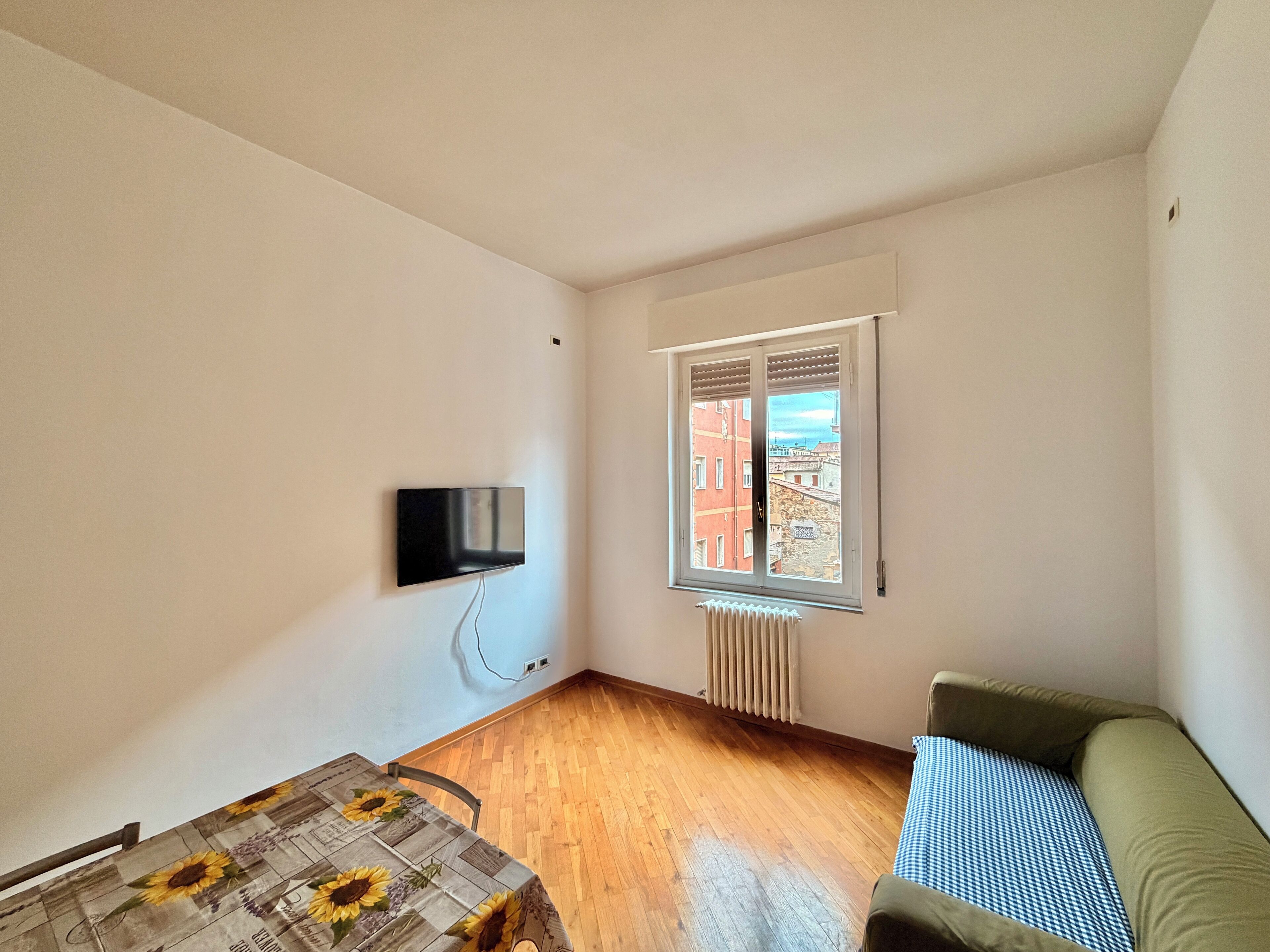 A room with a wooden floor, a wall-mounted TV, a window with a view of buildings, and a radiator.