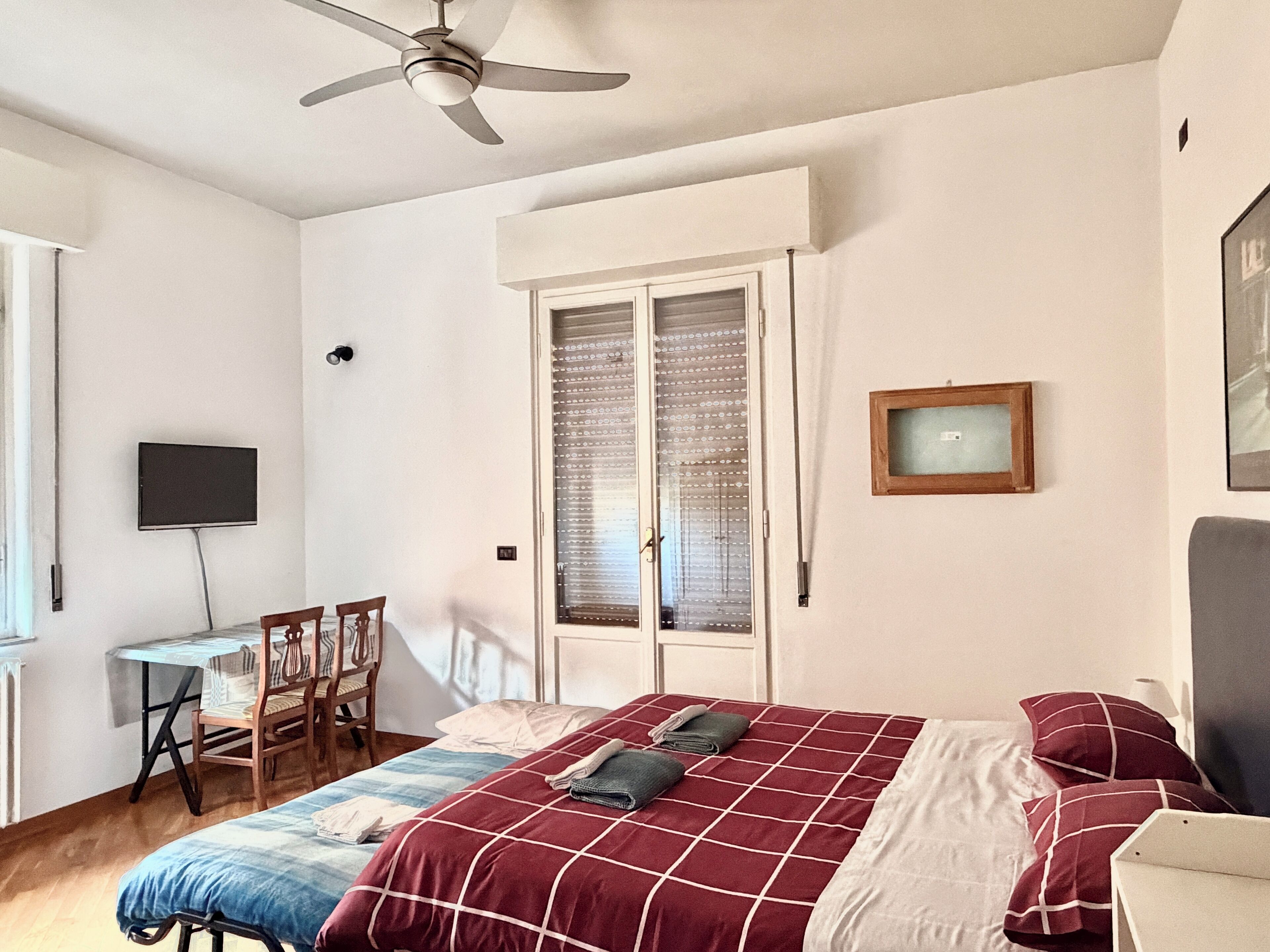 A bedroom with a bed, a desk, a chair, a television, and a window with blinds.