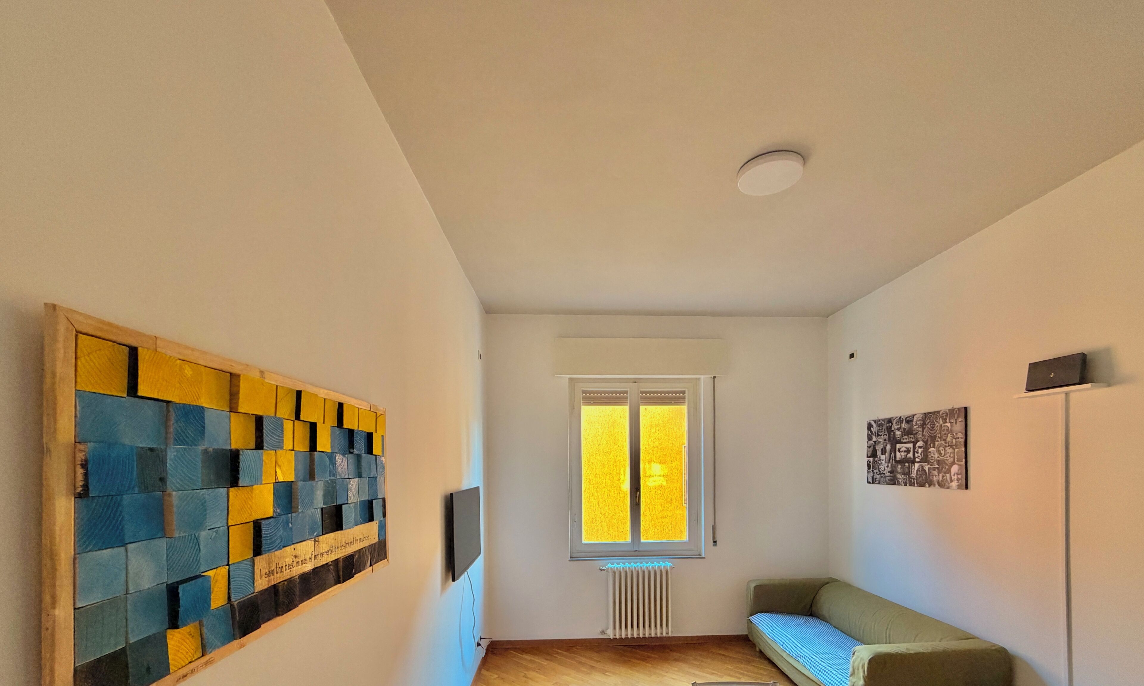 Un couloir avec un plancher en bois, une fenêtre avec des stores jaunes, un radiateur, un canapé vert et une œuvre d’art accrochée au mur.