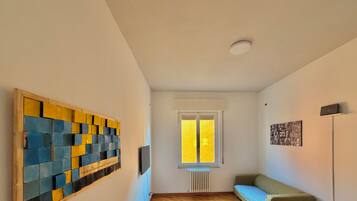 Un couloir avec un plancher en bois, une fenêtre avec des stores jaunes, un radiateur, un canapé vert et une œuvre d’art accrochée au mur.