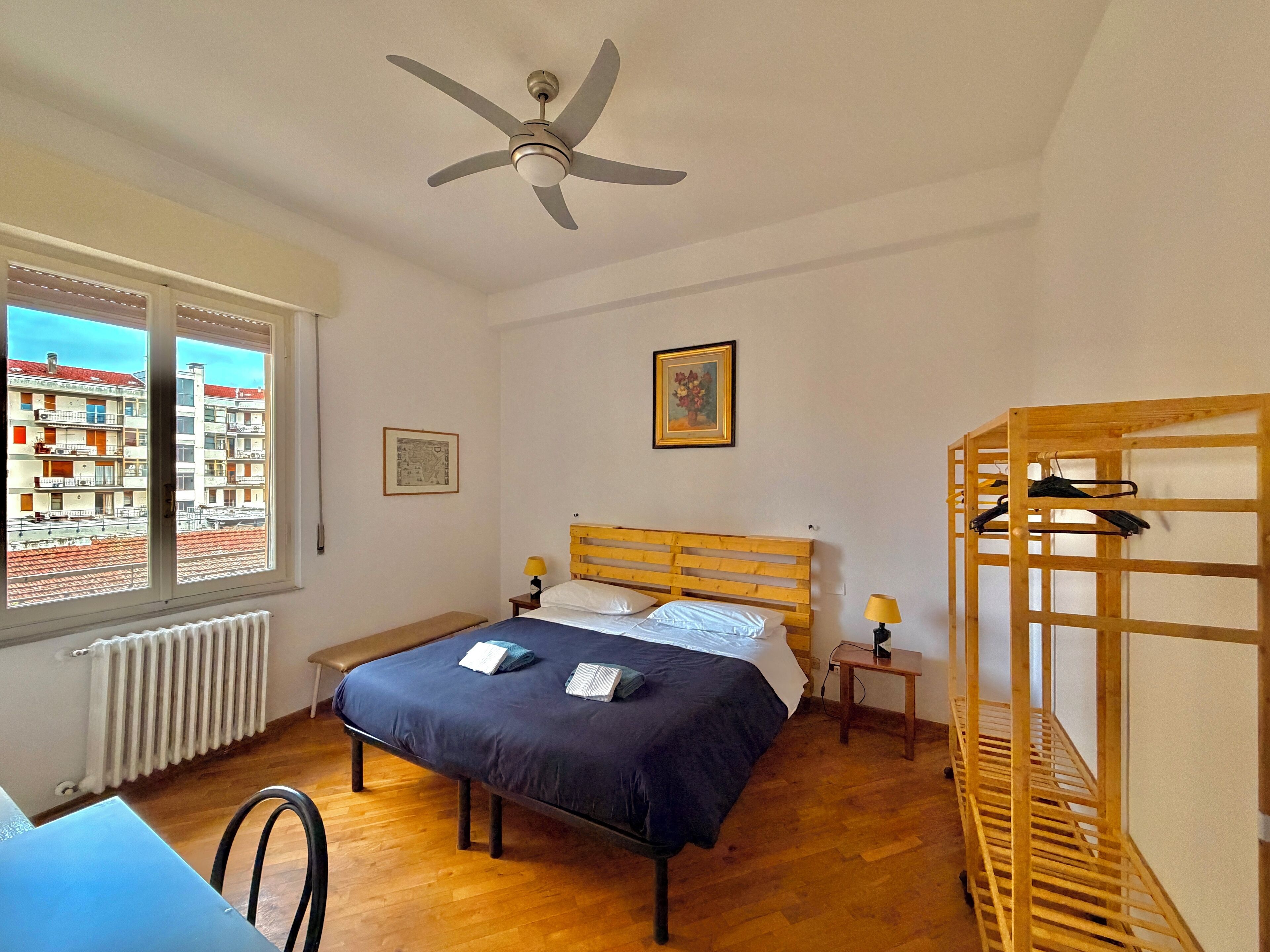 A bedroom with a bed, a wooden headboard, a ceiling fan, a window with a view of buildings, a radiator, a wooden shelf, and a small table with a lamp.