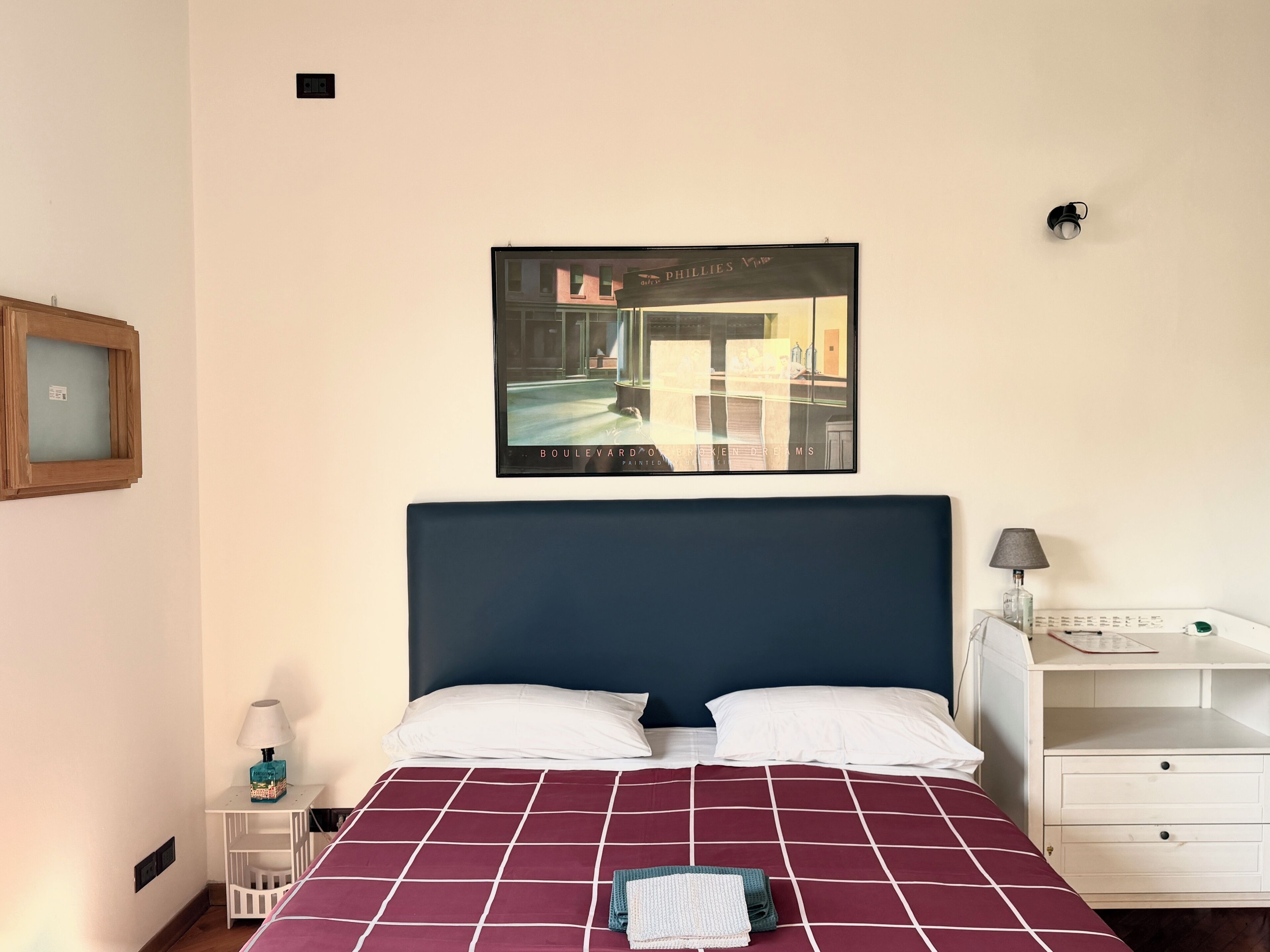 A hotel room with a large bed, a nightstand, a desk, and a wall-mounted picture.