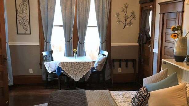 A bedroom with a large bed, a chandelier, a dining table with chairs, and a vase with flowers. - Three Danes Inn (Fort Worth)