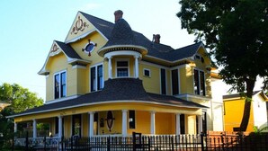 A yellow Victorian-style house with a wrap-around porch, a turret, and a black roof. - Three Danes Inn (Fort Worth)