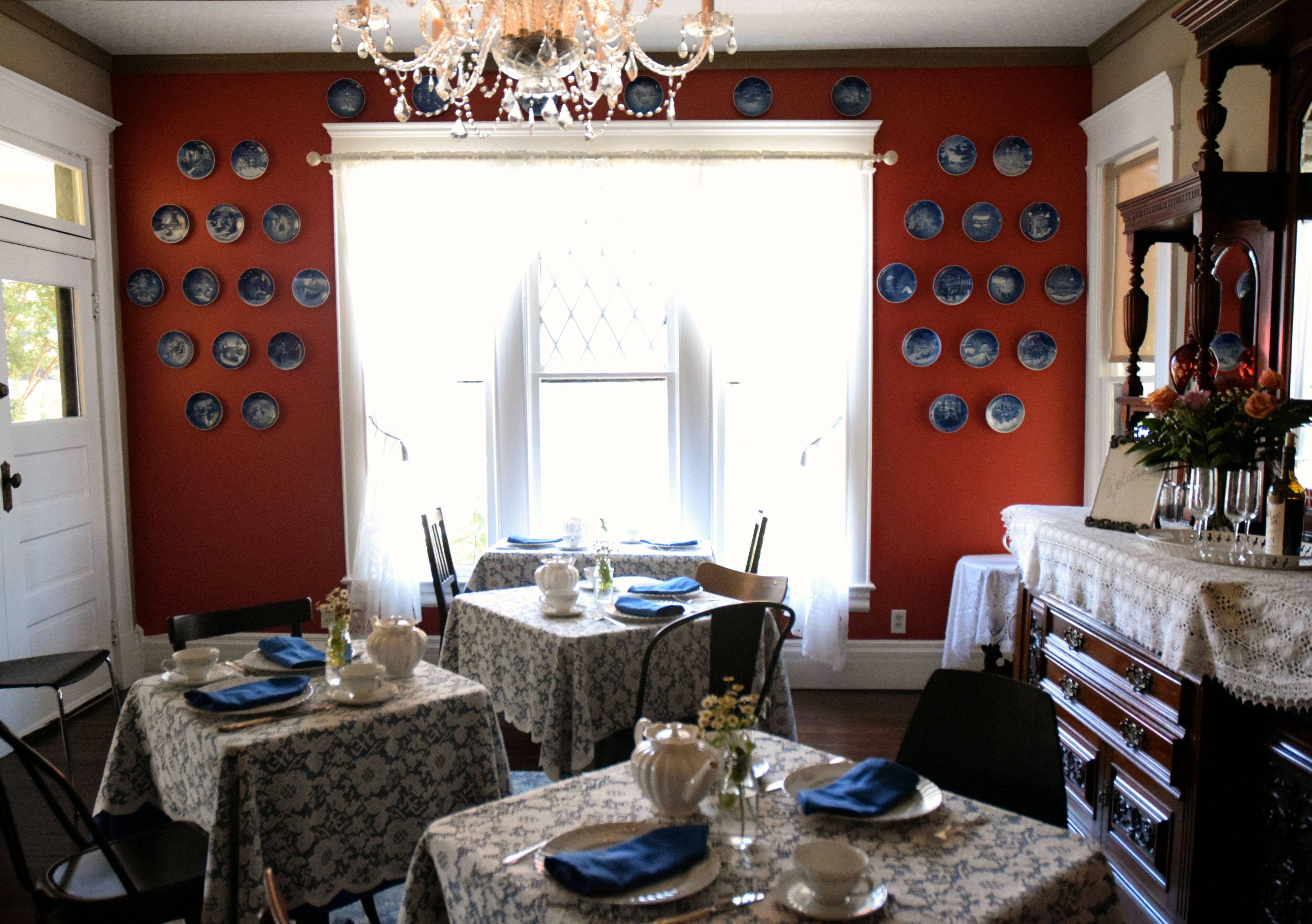 Ein Esszimmer mit vier gedeckten Tischen, einer Kronleuchter und einer roten Wand mit blauen Tellern.