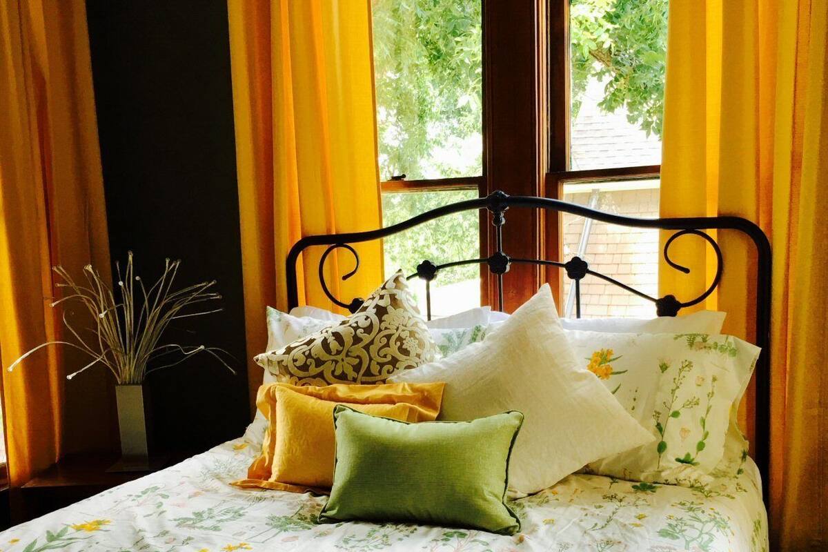 A bed with floral bedding, a fur throw, and multiple pillows in a room with yellow curtains and a window with greenery outside.