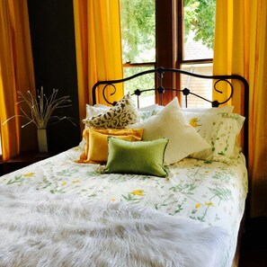 A bed with floral bedding, a fur throw, and multiple pillows in a room with yellow curtains and a window with greenery outside. - Three Danes Inn (Fort Worth)