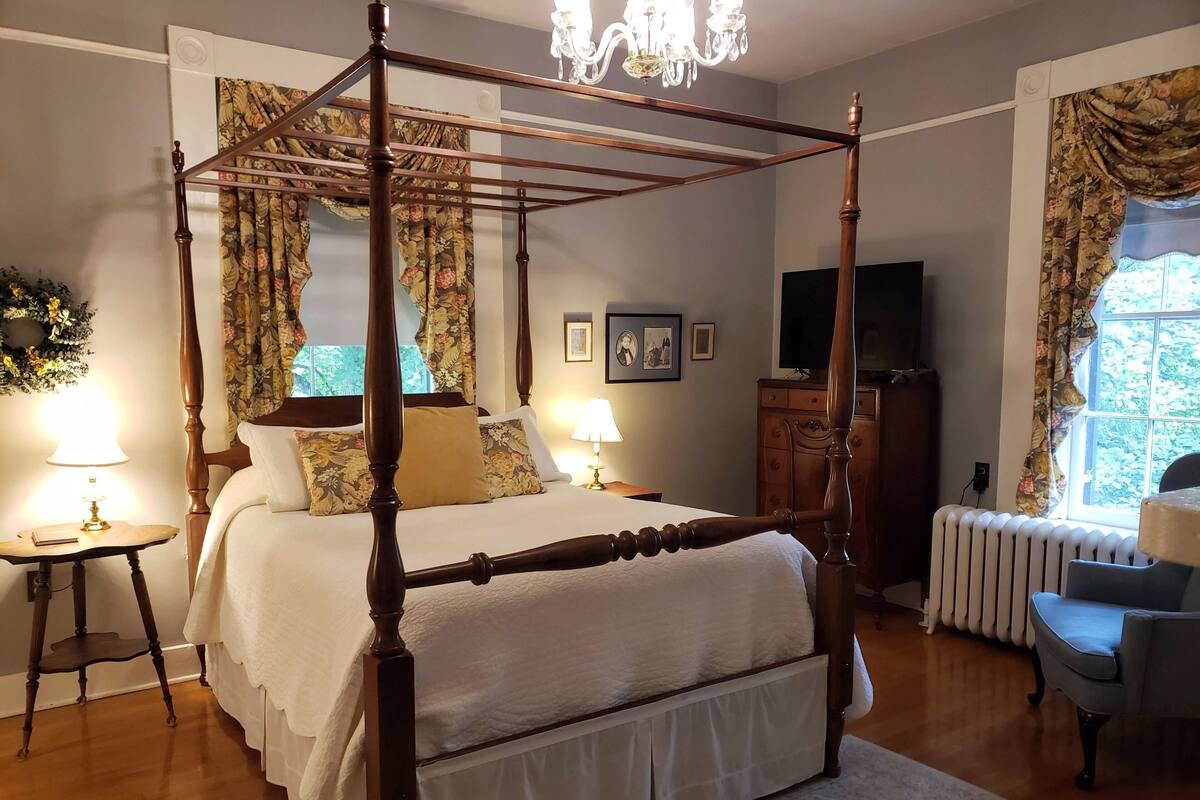 A four-poster bed with a white bedspread and multiple pillows, flanked by two bedside tables with lamps, a wooden dresser, and a radiator.