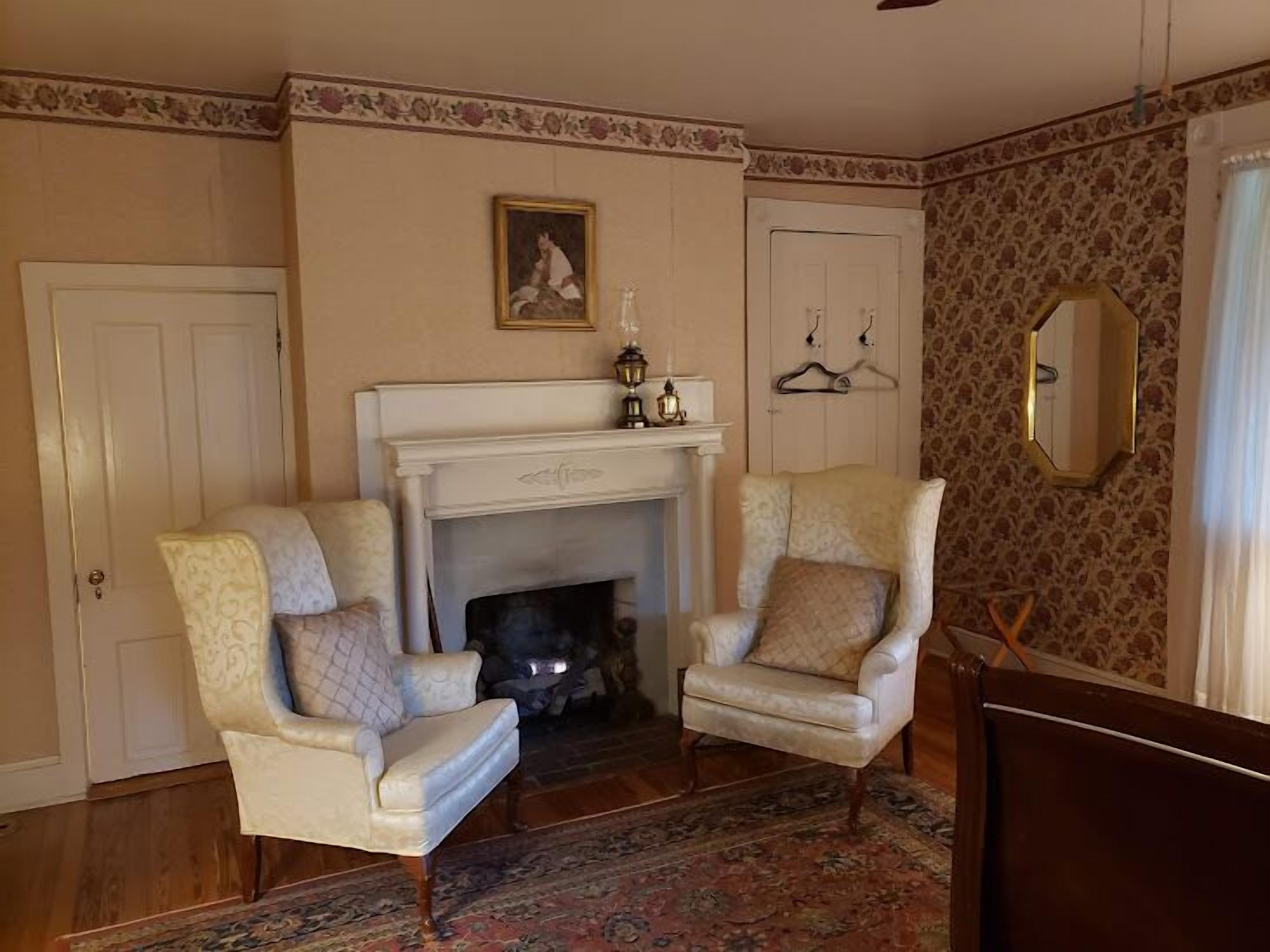Een kamer met een open haard, twee fauteuils, een gekleurd tapijt, een spiegel en een deur.