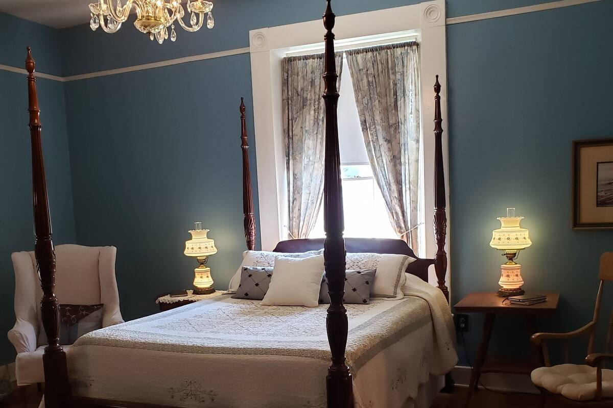 A four-poster bed with a canopy, two bedside lamps, a chair, and a small table with a lamp.