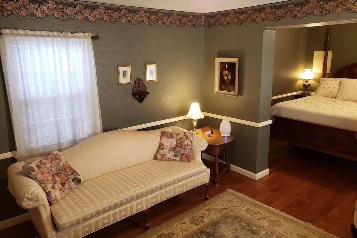 A cozy room with a sofa, a small table with a lamp, a bed, and a mirror.