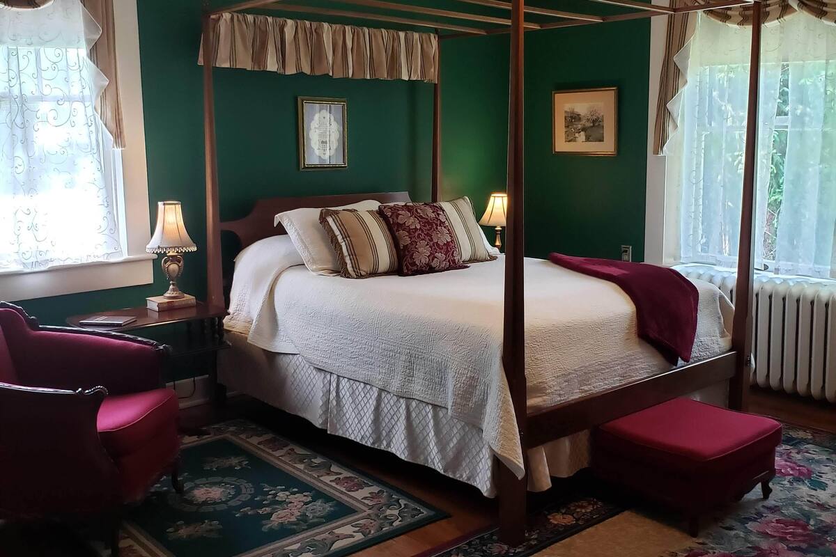 A four-poster bed with a white bedspread, flanked by two red armchairs, a wooden nightstand with a lamp, and a floral area rug.
