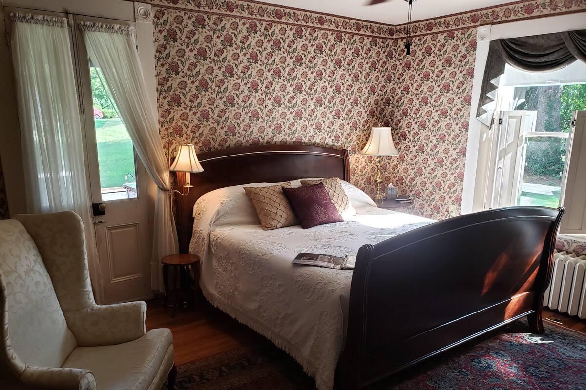 A bedroom with a bed, a chair, a nightstand, a lamp, and a window with curtains.