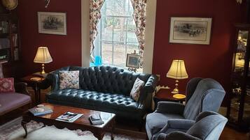 A living room with a dark blue sofa, a matching armchair, a coffee table, and a chandelier. There is a large window with floral curtains, a bookshelf, and two lamps.