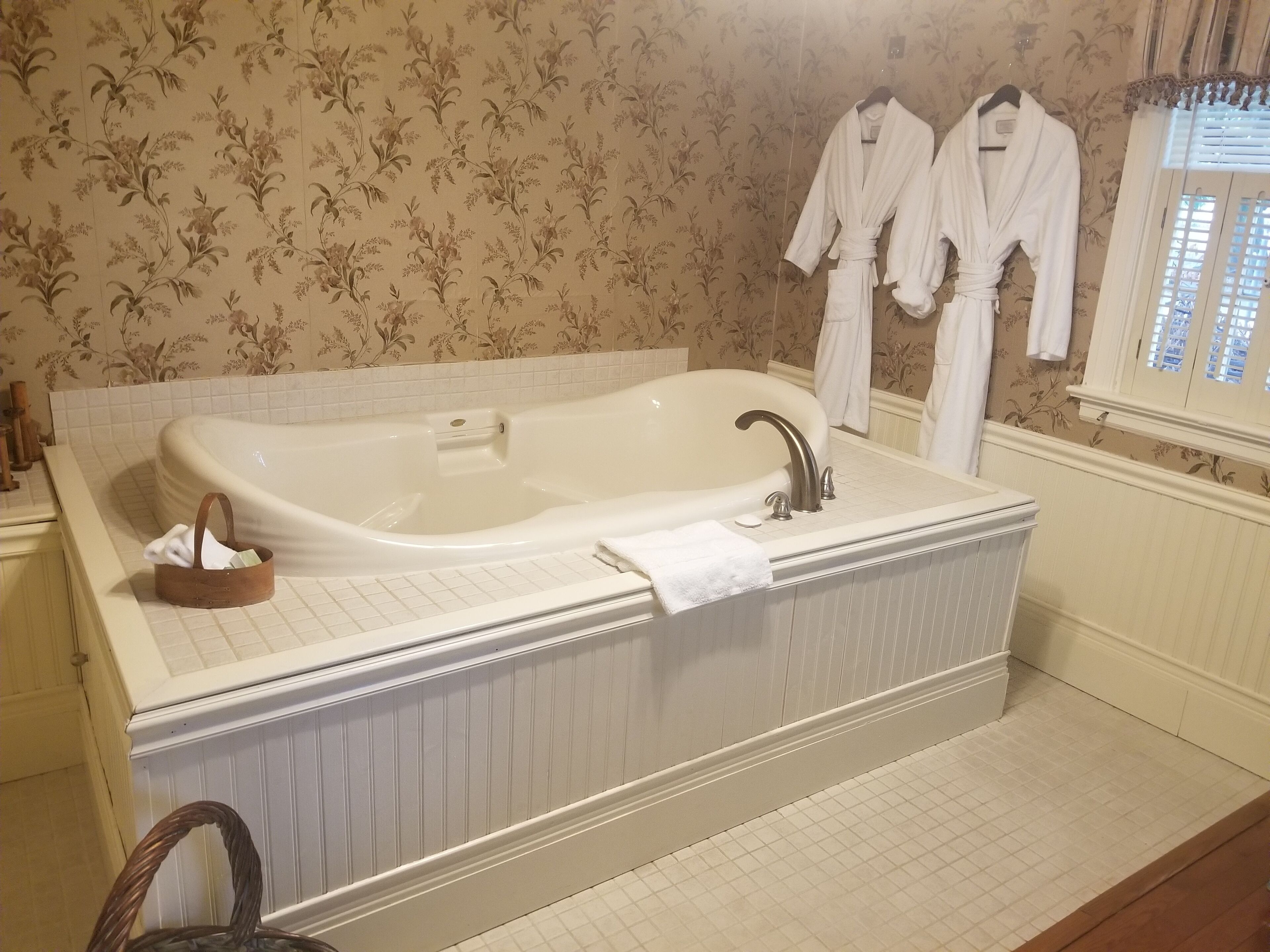 A bathroom with a large bathtub, a wooden basket, and two white robes hanging on the wall.