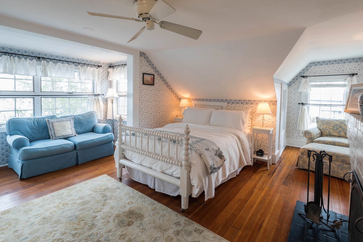 Ein Schlafzimmer mit einem Bett, einem blauen Sofa, einem weißen Sessel, einem Deckenventilator und einem Kamin.
