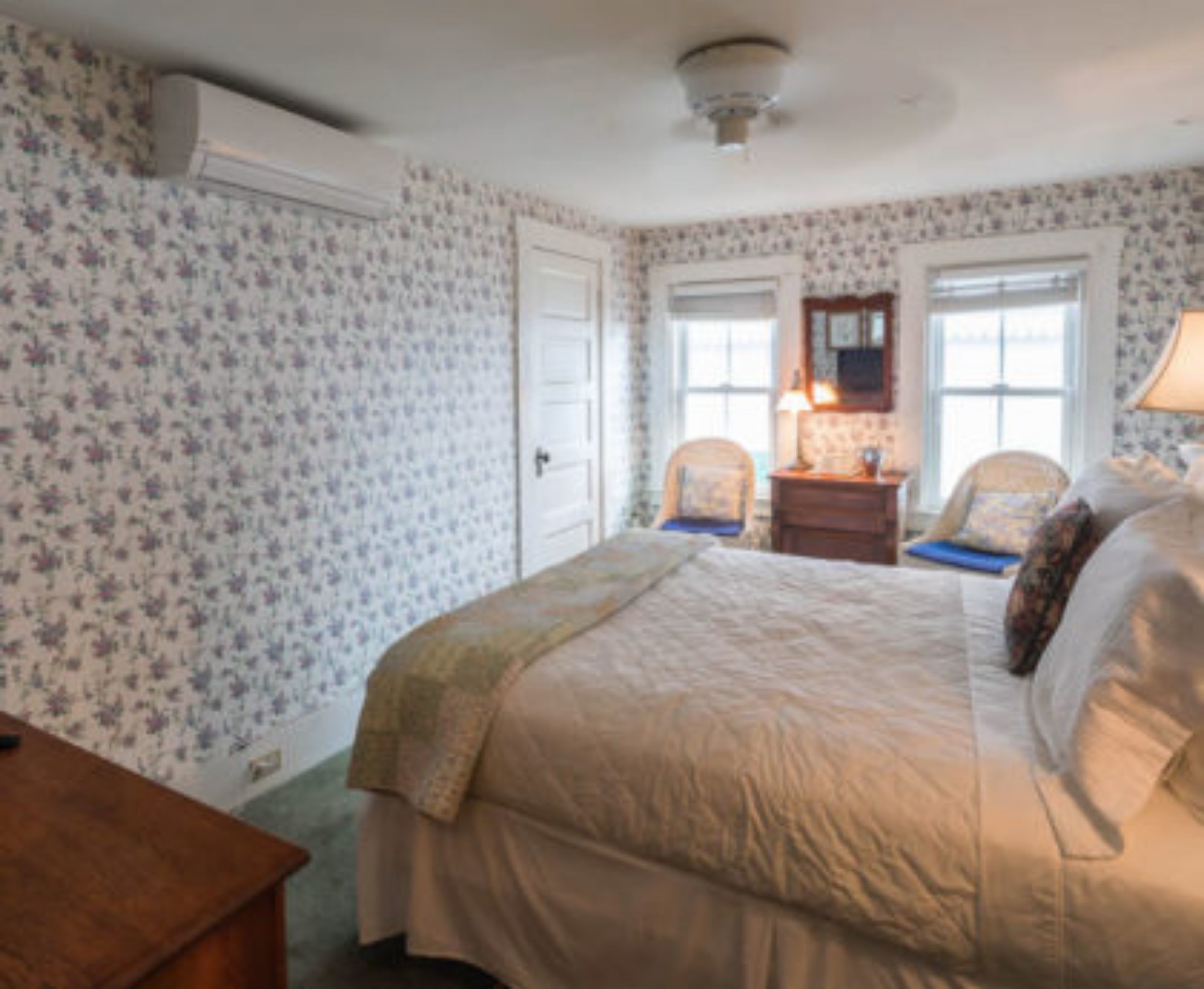 Une chambre à coucher avec un papier peint à motifs floraux, un lit recouvert d’une couvre-lit beige, deux chaises, une table de chevet en bois et une fenêtre donnant sur un paysage agréable.