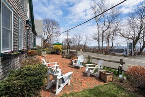 A patio with white Adirondack chairs, a brick pathway, and a view of a road and trees.