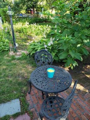 A garden patio with a table and chairs, surrounded by lush greenery and a brick pathway.
