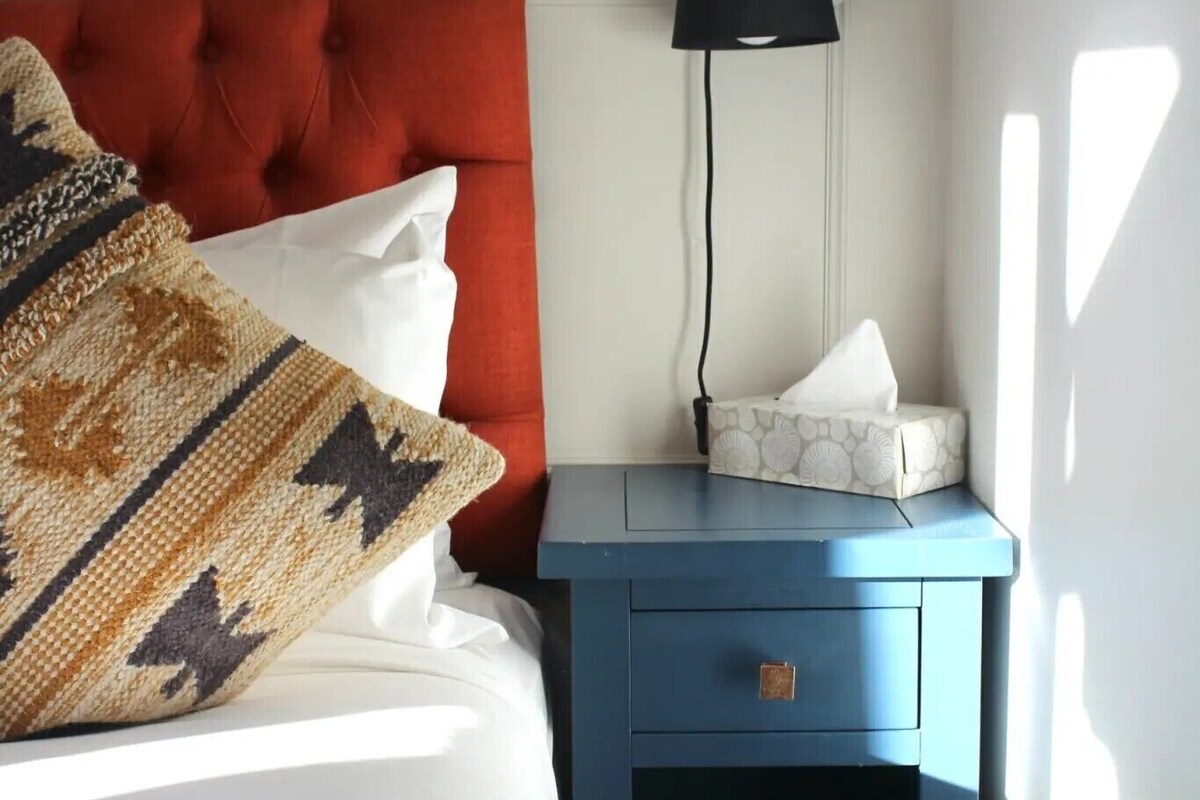 A bed with a red headboard, a patterned throw pillow, a white pillow, and a blue bedside table with a lamp and tissue box.