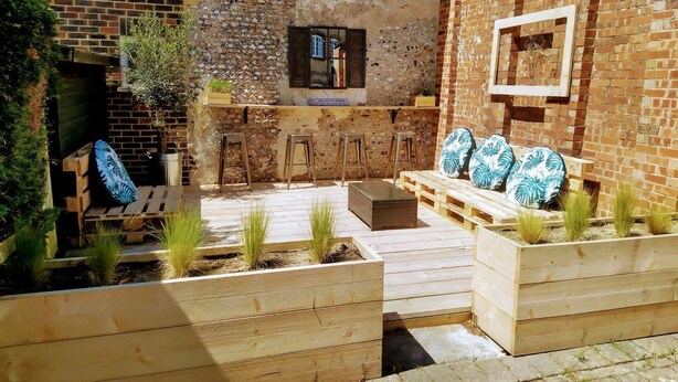 A rustic outdoor seating area with wooden benches and planters, a small bar area with stools, and a stone wall backdrop. - The Old Store Guest House (Chichester)