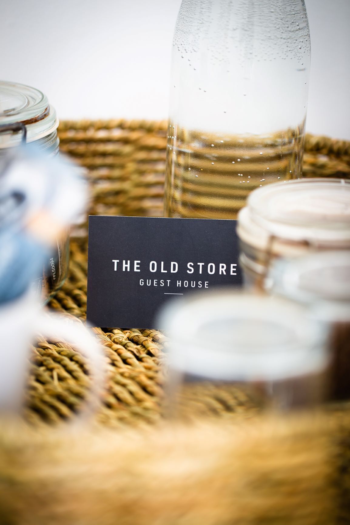 A black sign with white text reading "THE OLD STORE GUEST HOUSE" placed on a woven surface with jars and a glass bottle.