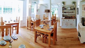 A spacious dining area with wooden tables and chairs, a kitchen with white cabinets, and a bright, naturally lit environment. - The Old Store Guest House (Chichester)