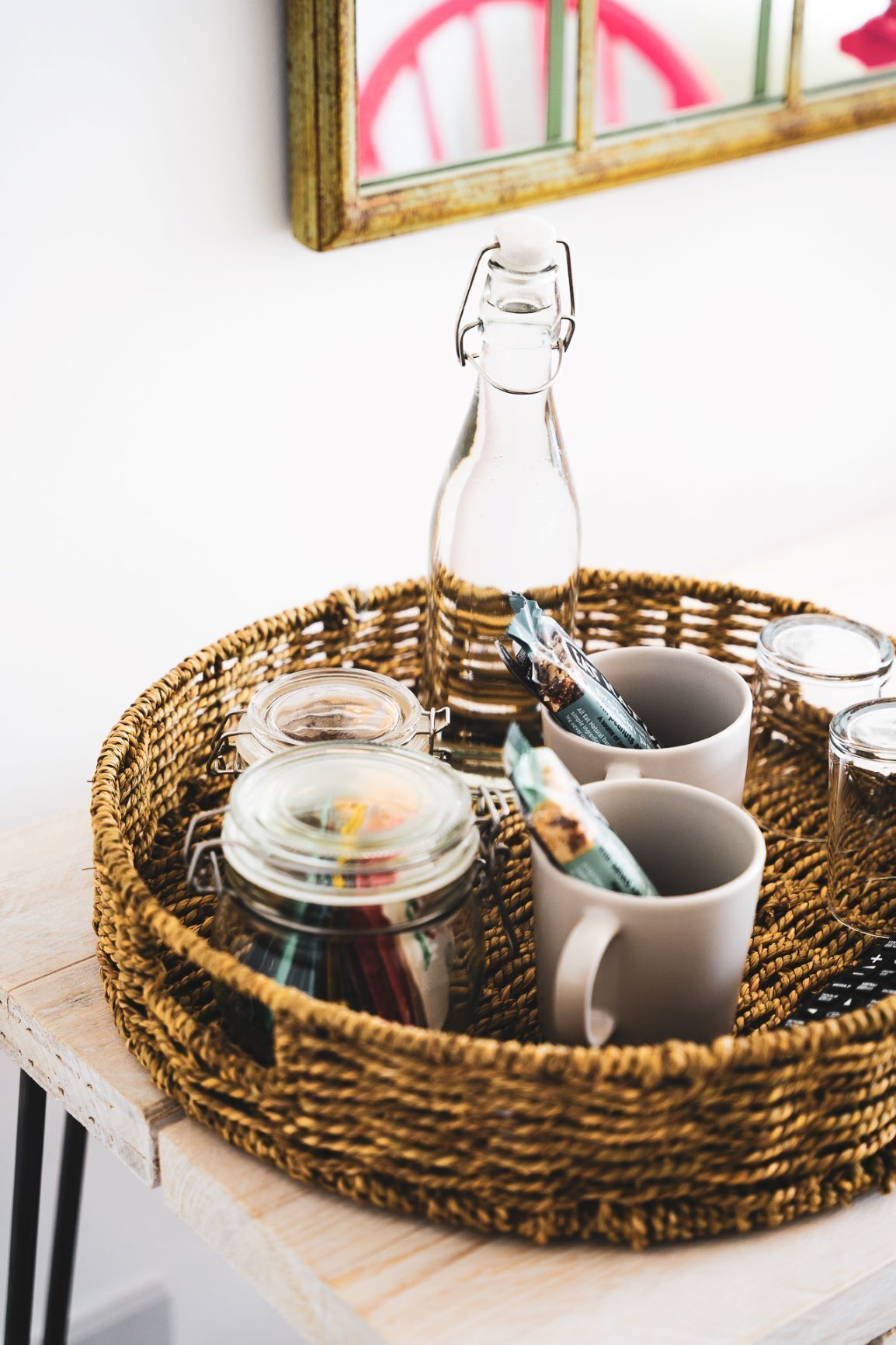 A wicker tray with a glass bottle, a jar, and two mugs on a wooden surface.