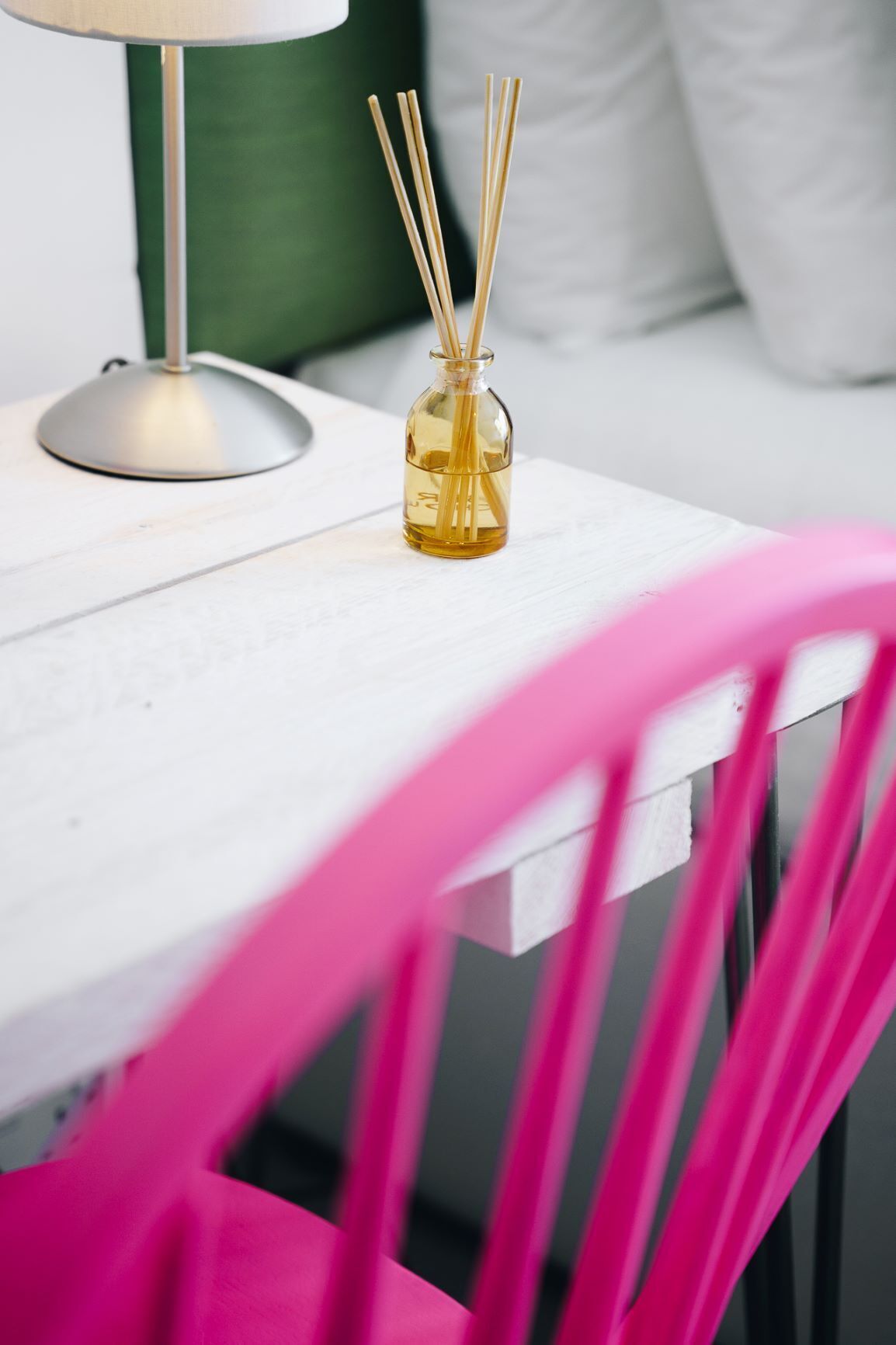 Ein weißer Tisch mit einer Glasflasche, gefüllt mit Schilfrohhröllchen, einer Lampe und einem grünen Kissen im Hintergrund.