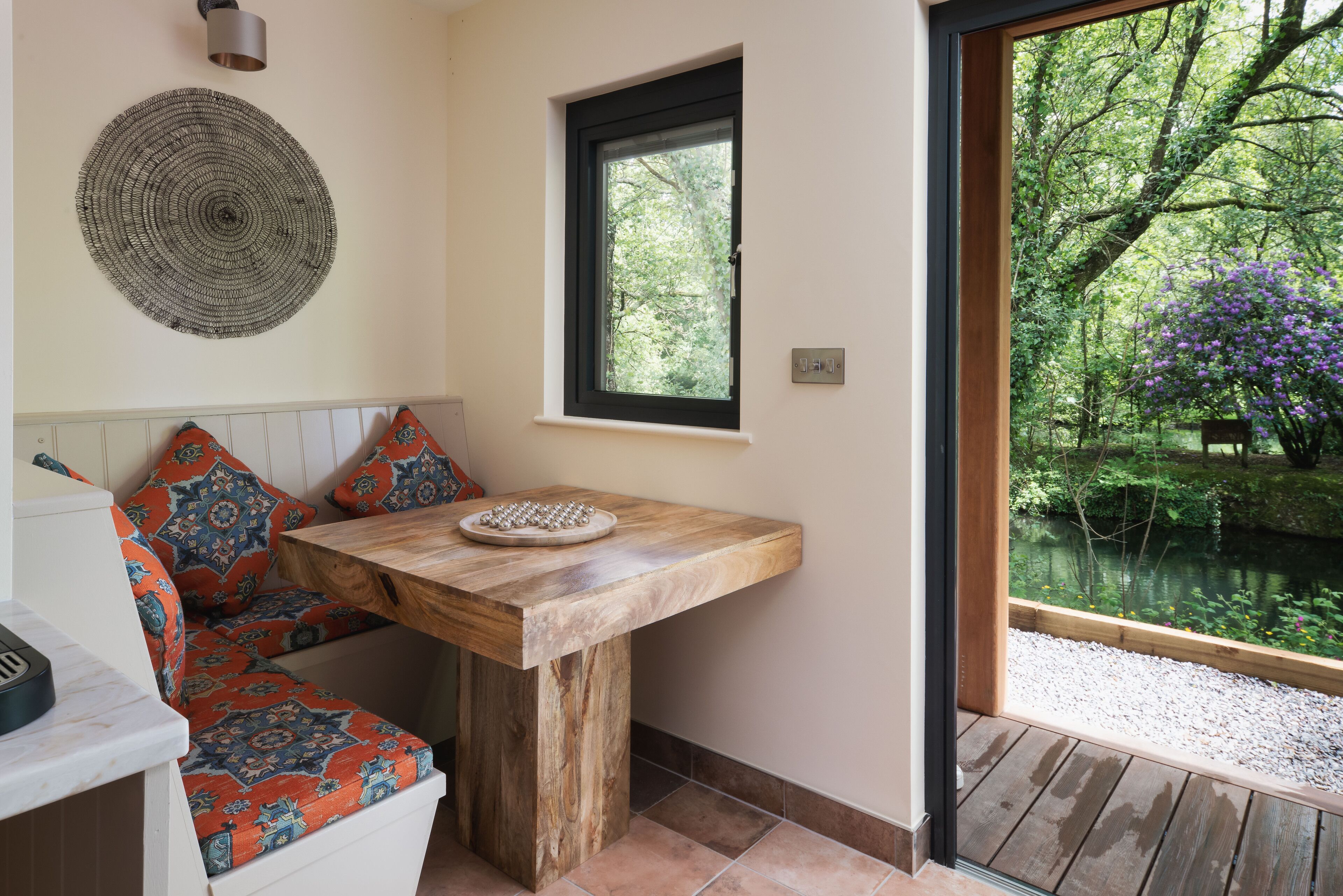 Ein Zimmer mit Holztisch, einer Bank mit gemusterten Kissen, einem Wandbehang aus Bändern und einem Fenster mit Blick auf Grünflächen und einen Teich.