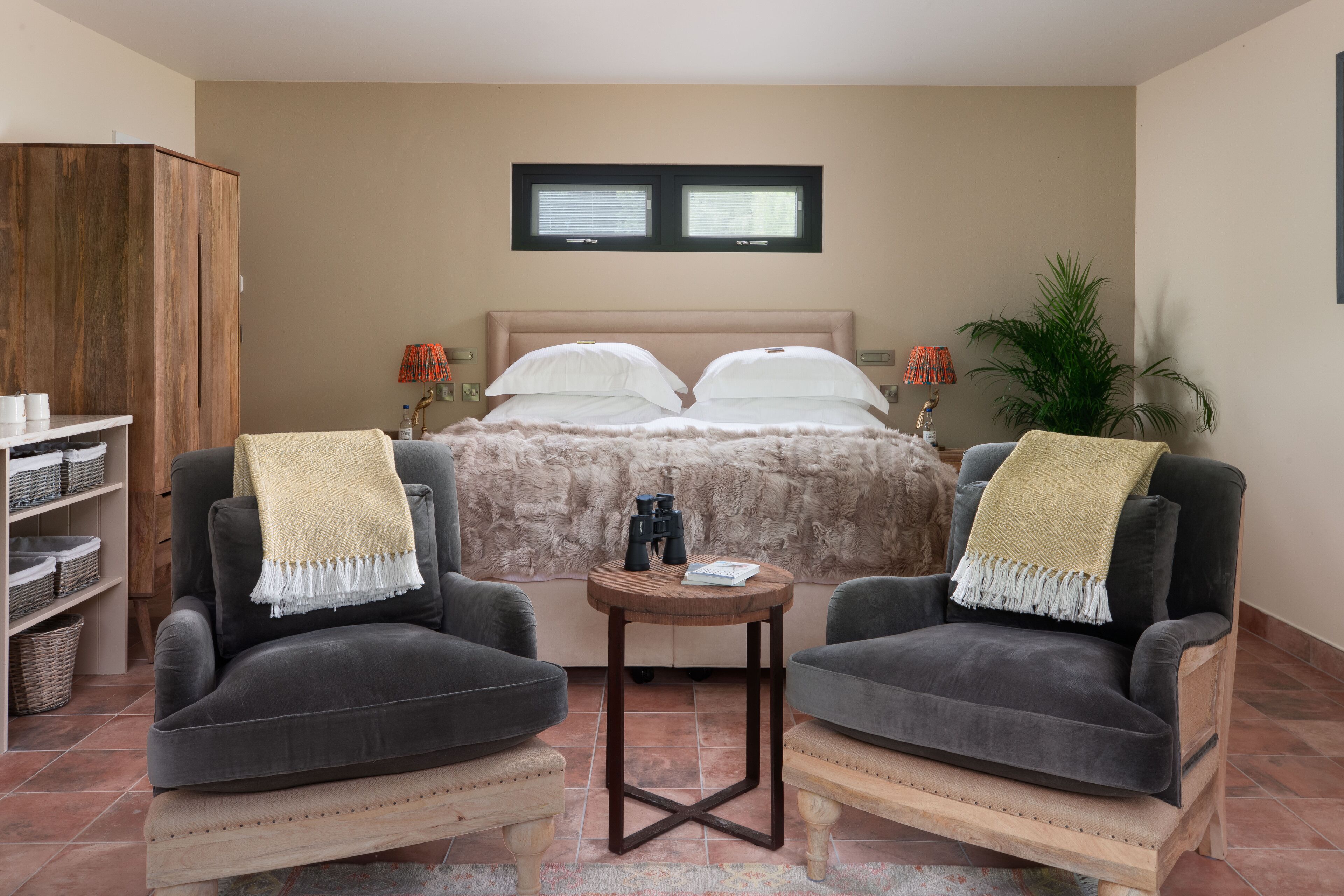 Une chambre à coucher comprenant un lit, deux fauteuils, une petite table ronde, une bibliothèque et une fenêtre avec des stores.