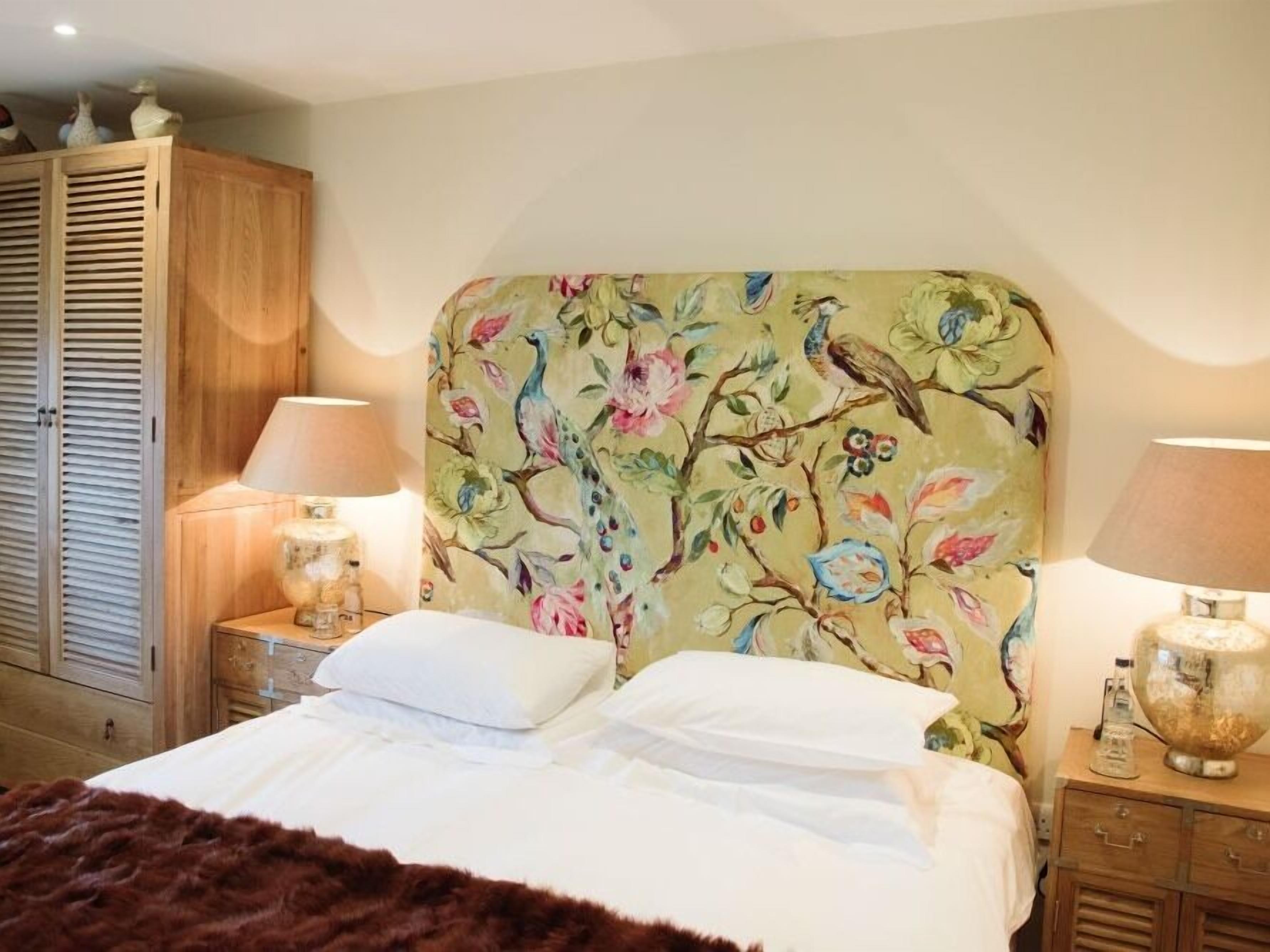 A bedroom with a floral headboard, a bed with white linens, two bedside lamps, a wooden wardrobe, and a brown fur throw.