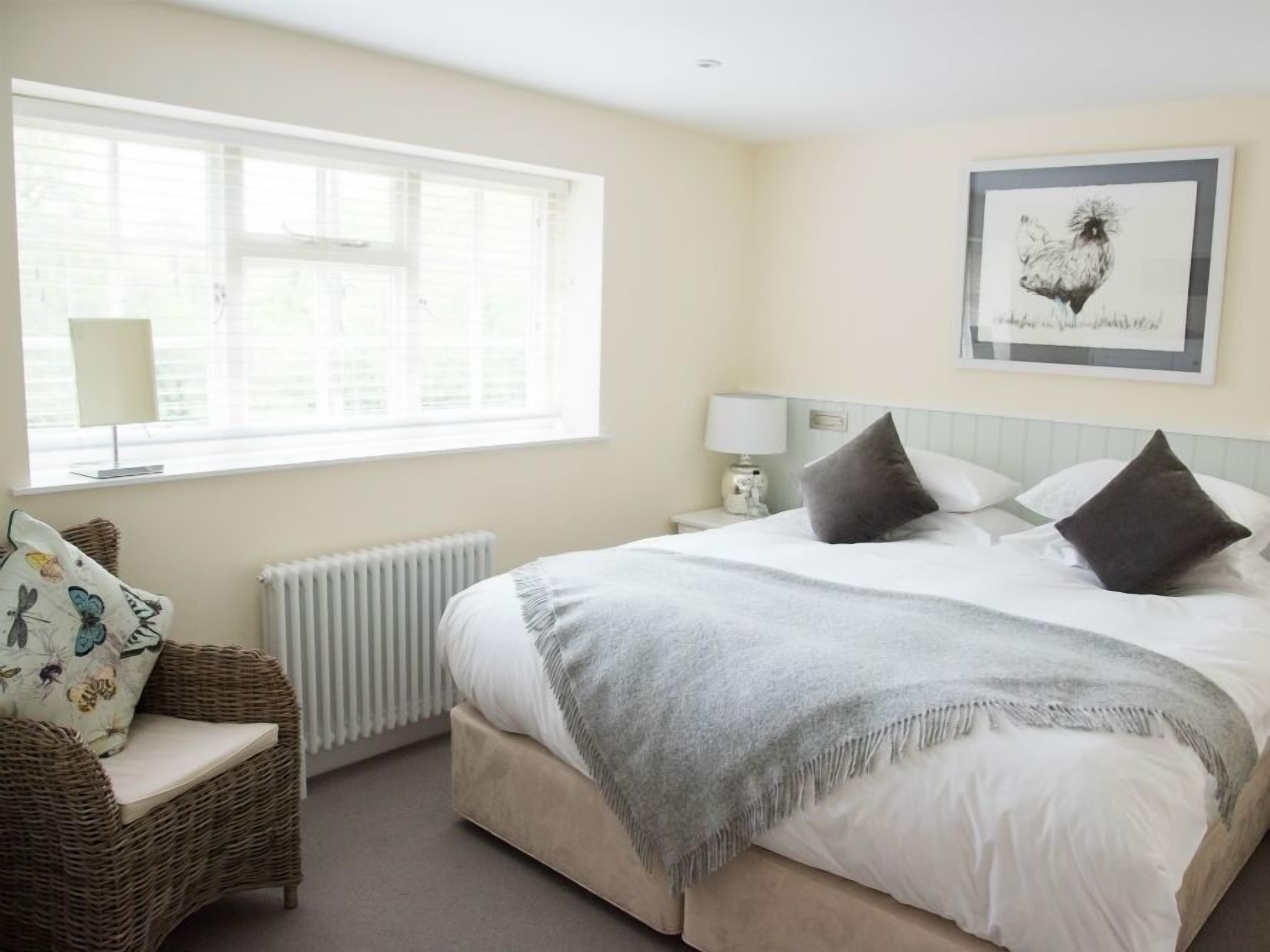 Une chambre avec un grand lit, un radiateur, une chaise en osier, une table de chevet avec une lampe et un tableau encadré au mur.