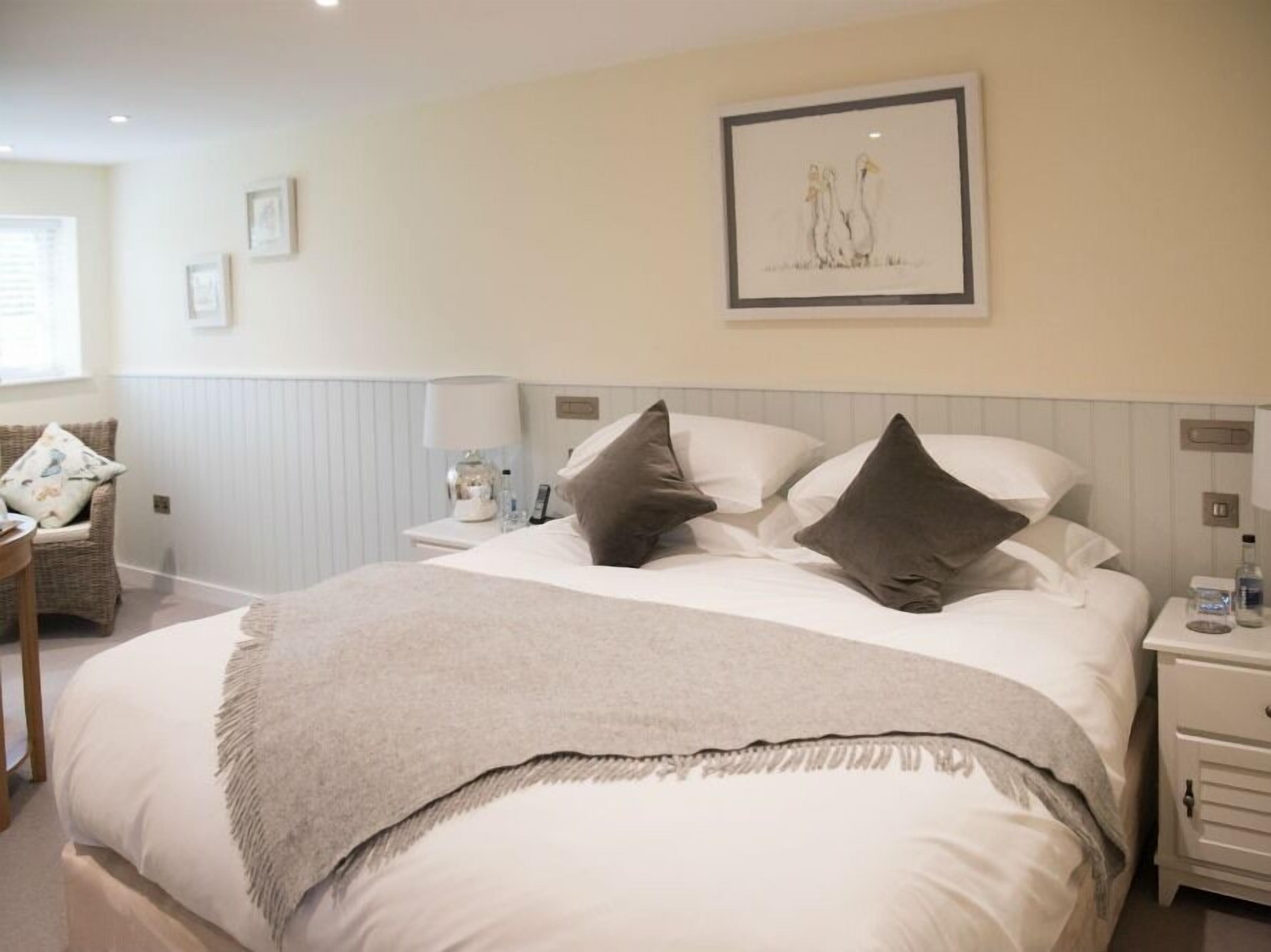 A neatly arranged hotel room with a large bed, a bedside table with a lamp, a chair with cushions, and a framed picture on the wall.