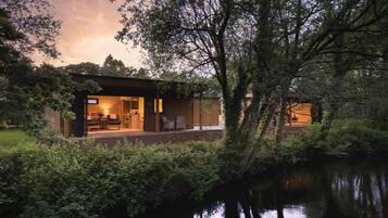 A modern house with a glass door leading to a dining area, surrounded by lush greenery and a calm water body.