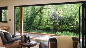 A room with a large window overlooking a garden with a pond and trees. There is a sofa and a small table with a kettle and cups. - The Turtley Corn Mill (South Brent)