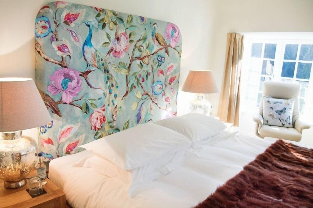 Un dormitorio con cabecera de flores, una cama con ropa de cama blanca, un edredón marrón, una mesita de noche con una lámpara, una silla con estampado de mariposas y una ventana con cortinas.